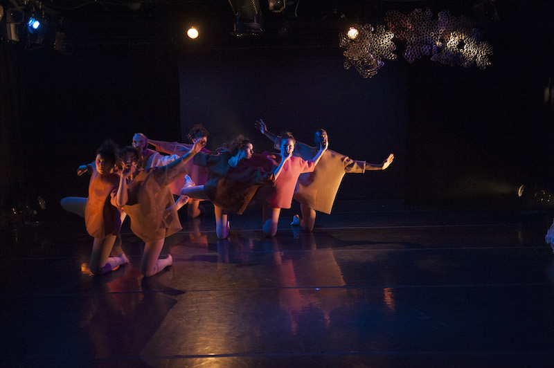 A group of dancers in pink and mustard hued boxy shifts kneel under an artful metal installation